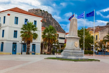 A marble monument in Nafplio square with palm trees and Palamidi Fortress on the hill.