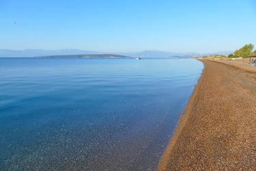 Calm clear blue sea meeting a pebble beach shoreline with distant mountains under a bright sky.