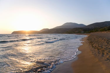 Golden sunset over a sandy beach with ocean waves and mountain landscape in the background.
