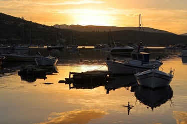 Small fishing boats anchored in a calm bay at sunset with mountain silhouettes in the background.