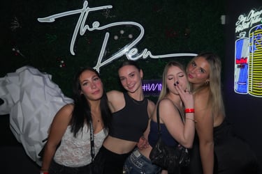 Four young women posing together at a nightclub under a white neon The Den sign.
