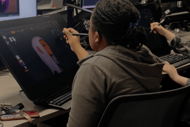 a creative student sitting at a desk with two monitors and a laptop