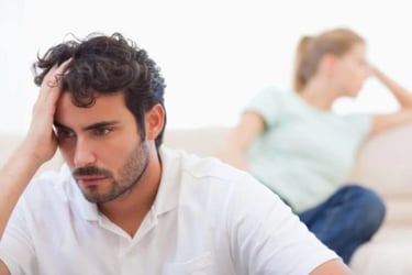 A stressed man holding his head during a relationship conflict with his partner sitting blurred in the background.