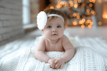 Baby girl with white flower headband on a white knit blanket with Christmas lights bokeh background.