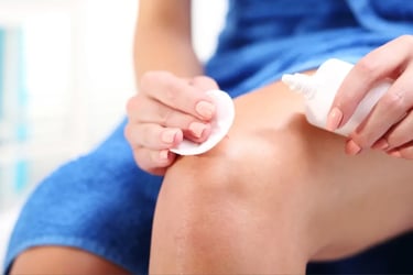 A woman applies topical skin cream to her knee using a cotton pad for targeted skincare treatment.