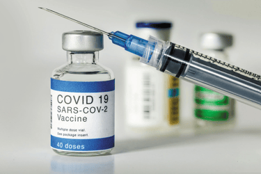 A medical syringe leaning against a COVID-19 SARS-CoV-2 vaccine vial on a white surface.