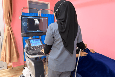 A healthcare professional in a hijab performing a diagnostic ultrasound on a patient in a pink medical room.