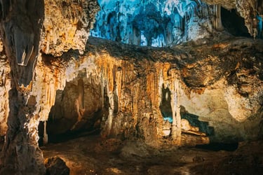 Illuminated underground limestone cave with ancient stalactites and stalagmites in varying colors.