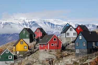Colorful coastal houses in a Greenlandic village with snowy mountain peaks and clouds in the background.