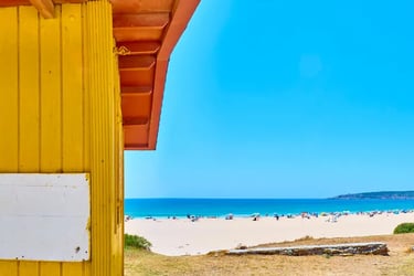 Yellow wooden beach hut overlooking a sunny white sand beach with turquoise ocean water and people.