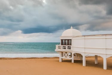 A white pavilion on La Caleta beach in Cadiz, Spain under a dramatic cloudy sky.