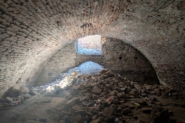 Sunlight streams through a stone opening into an ancient vaulted brick cellar with rubble in Nis