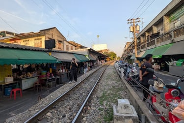 A narrow railway track runs through the Tala Market in Thailand with shops and stalls on both sides.
