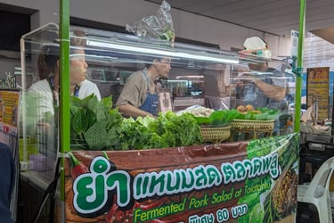 Street food vendor in Thailand serving fermented pork salad with fresh greens and vegetables at a night market.