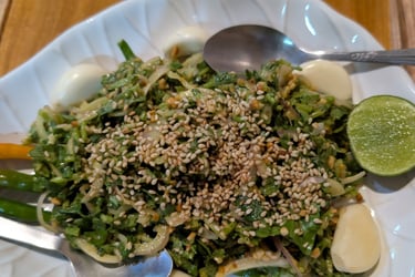 A fresh Burmese pe salad topped with toasted sesame seeds, garlic cloves, and lime on a white plate.