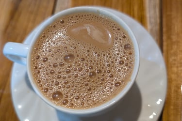 A cup of hot frothy Burmese sweet tea on a white saucer on a wooden table.