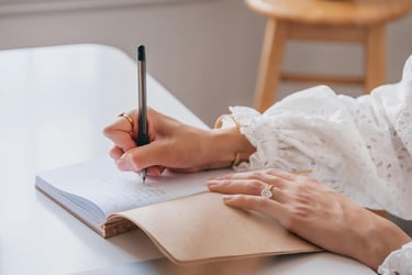 Ein aufgeschlagenes Notizbuch auf einem Tisch. Eine Frau schreibt mit dem Stift etwas in das Buch.