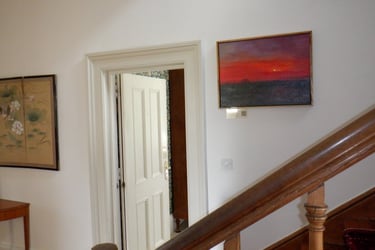 Oil painting of a sunset on a gallery wall near a wooden staircase and white doorway.