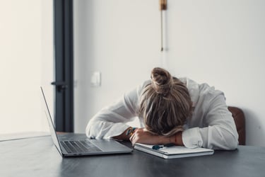 An exhausted woman napping on a notebook next to a laptop, illustrating burnout and work fatigue.