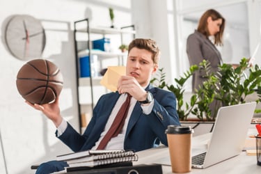 Confused businessman holding a basketball and sticky notes at his office desk with a laptop.