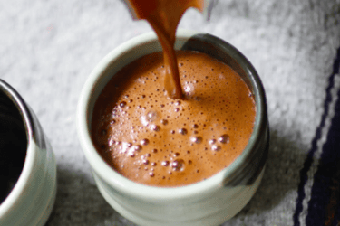 Rich hot chocolate being poured into a ceramic mug with frothy bubbles on the surface.