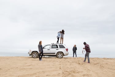 A group of friend near a beach front with a car parked in the middle enjoying their day and giggling