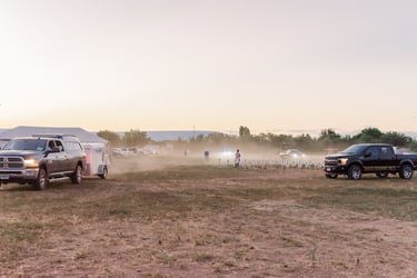 Trucks and trailers parked in a dusty field during an outdoor sunrise event with people gathered.