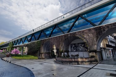 Modern glass pedestrian bridge spanning over a historic stone archway in a European city plaza.