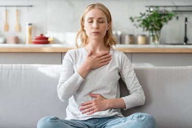 A blonde woman practices deep breathing and mindfulness while sitting on a sofa in a modern kitchen.