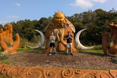 Lloyd & Adam posing in front of a giant sun and moon goddess sculpture at an outdoor art park.