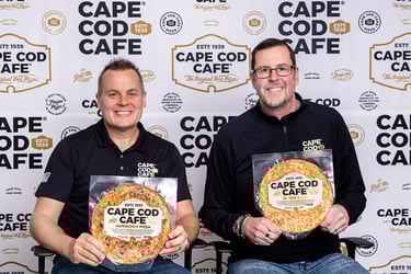 Two men hold Cape Cod Cafe frozen bar pizzas in front of a company logo step and repeat backdrop.