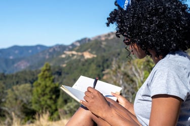 A woman with curly hair writes in a journal while sitting outdoors with a scenic mountain view.
