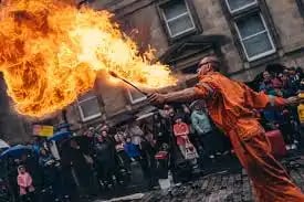 A fire breather in an orange jumpsuit performs a spectacular street show for a crowd.