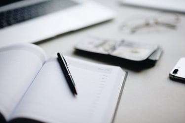 A black pen resting on an open paper planner next to a stack of cash and a laptop on a white desk.