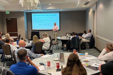 A presenter leads a business seminar on virtual success and shared support models for a group of professionals seated at