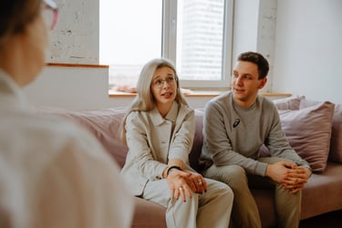 A young couple sitting on a pink sofa in a therapy session discussing relationship issues with a counselor.