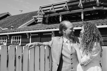 Bride and groom laughing together while leaning on a white wooden fence.