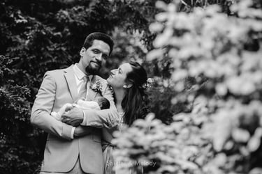 Bride and groom standing among trees with a baby.