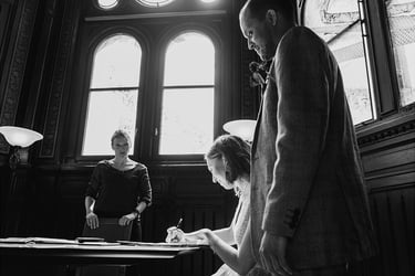 Bride signing marriage documents with groom looking over her shoulder.