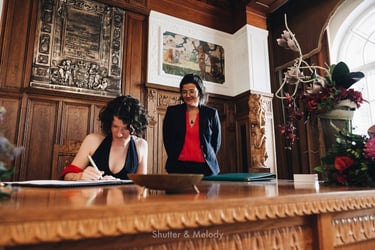 Die Braut unterschreibt die Heiratsurkunde, während ihre Frau ihr über die Schulter schaut.