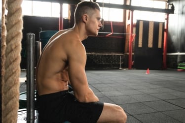 Shirtless muscular man resting between sets during a fitness workout at a cross-training gym.