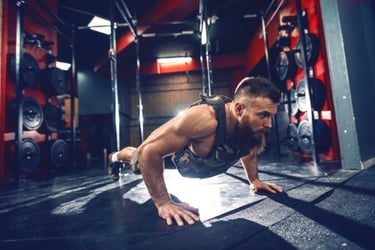 Muscular bearded man performing weighted pushups in a camouflage vest at a modern gym.