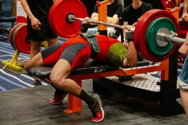 A powerlifter in a red singlet performs a heavy bench press with weighted plates at a gym competition.