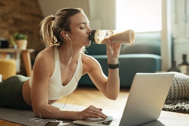 A woman drinking a protein shake while using her laptop during a home workout session.