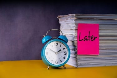 A blue alarm clock next to a stack of paperwork with a pink sticky note that says Later, symbolizing procrastination.