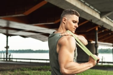 Athletic man performing a shoulder stretch using a green resistance band outdoors.