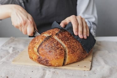 A person slicing a fresh loaf of crusty homemade sourdough bread on a wooden board.