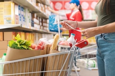 A person checking a grocery list next to a full shopping cart with paper bags in a supermarket aisle.