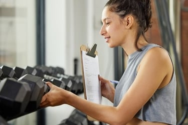 A female personal trainer holding a clipboard while organizing dumbbells on a gym weight rack.