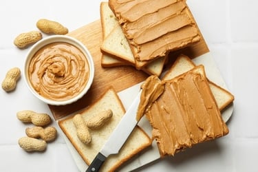 Creamy peanut butter spread on slices of white bread with whole peanuts on a wooden cutting board.